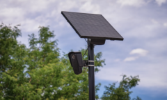 A Flock camera installed by the company Flock Safety who's office is located in Georgia. A brown metal pole installed vertically into the ground holds up a oval like housing with a surveillance camera mounted inside and above that is a square solar panel that is providing power for the camera to work