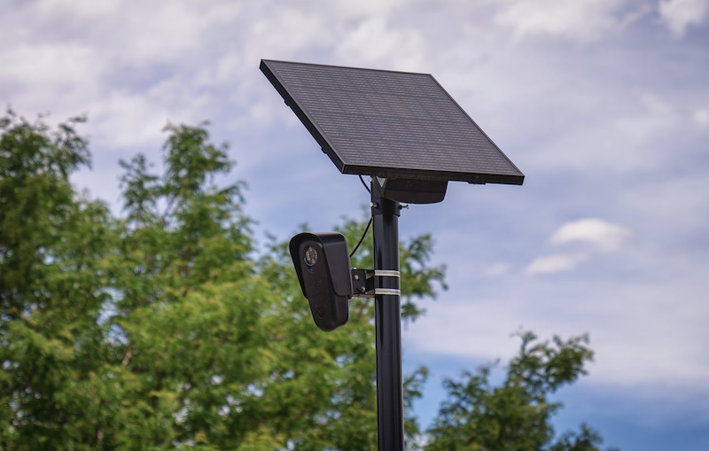 A Flock camera installed by the company Flock Safety who's office is located in Georgia. A brown metal pole installed vertically into the ground holds up a oval like housing with a surveillance camera mounted inside and above that is a square solar panel that is providing power for the camera to work