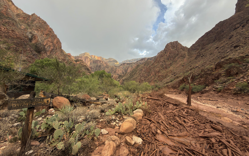 A picture of what remains after major flooding inside the Crand Canyon and the Colorado River. There is a National Park Service wooden post that is severely damaged and the landscape show mud, rocks, debris and vegetation all forcefully pushed downstream.